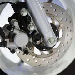 Primer plano del freno delantero de una scooter: pinza plateada, disco perforado y horquilla.