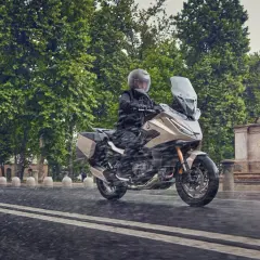 Motociclista en moto beige con alforjas, rodando bajo la lluvia. ¿Cuántos kilómetros dura una moto 125? ¡Esta parece lista para un largo viaje!