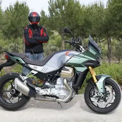 Motociclista con casco posa junto a una moto Guzzi Mandello verde y plateada, lista para la aventura en un camino rural.