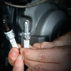 Manos sostienen dos bujías, una sucia y otra nueva, para una scooter.