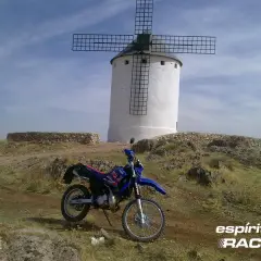 Yamaha DT 125 RE azul y roja aparcada junto a un molino de viento blanco en un paisaje rocoso.