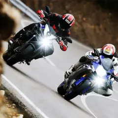 Dos pilotos compiten en una carretera sinuosa, uno en una Yamaha mas rápida, inclinándose en una curva cerrada.