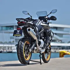 Voge 900 DSX ficha técnica: una moto de aventura negra con detalles dorados, aparcada junto a un puerto con veleros y un edificio moderno.
