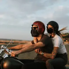 Pareja con mascarillas, montando a horcajadas moto al atardecer. El viento agita el cabello de ella.