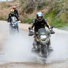 Motociclistas en un **conti trail attack 4 test** atraviesan un charco de agua en un camino de tierra, levantando una gran estela.