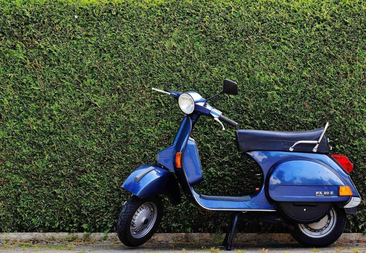 Azul Vespa PX 80 E, un clásico que evoca los modelos Vespa años 60, aparcada frente a un seto verde.