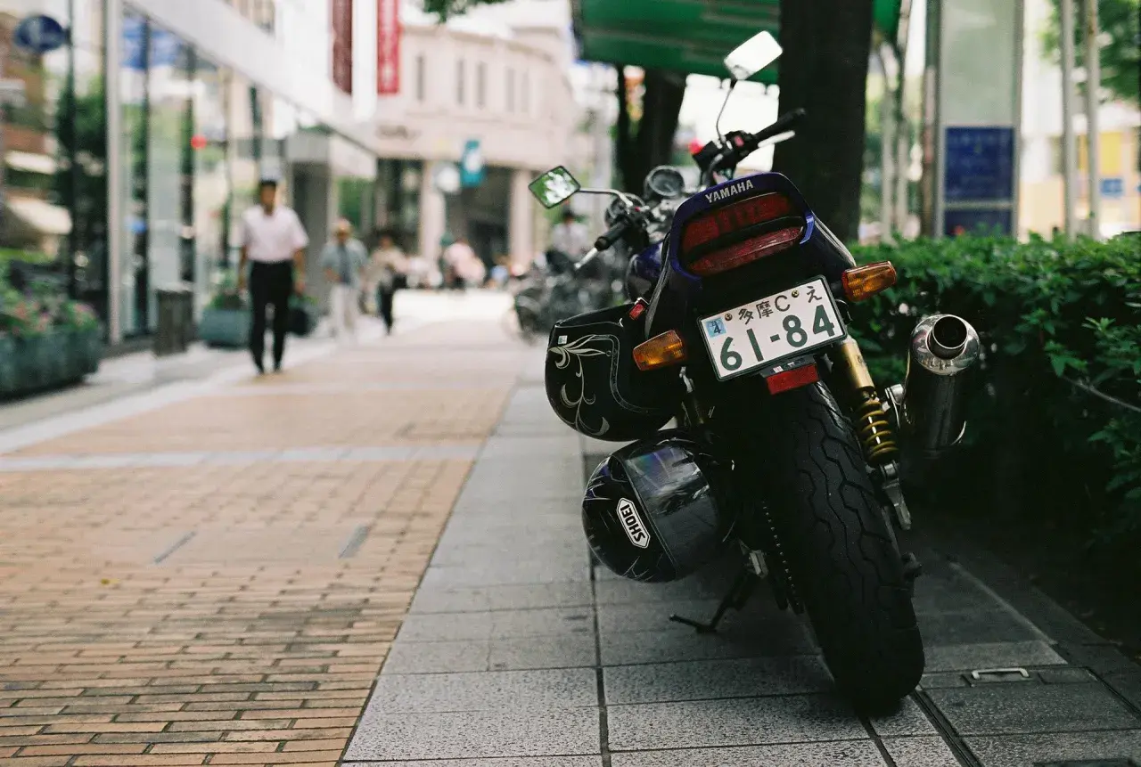 Una moto Yamaha aparcada en la acera, con dos cascos colgados. Al fondo, gente pasea por la calle.