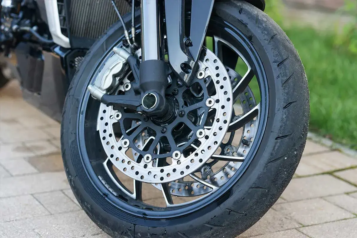 Primer plano de la rueda delantera de una motocicleta, con discos de freno perforados y pinzas. Se ve el neumático desgastado, listo para reparar rueda.