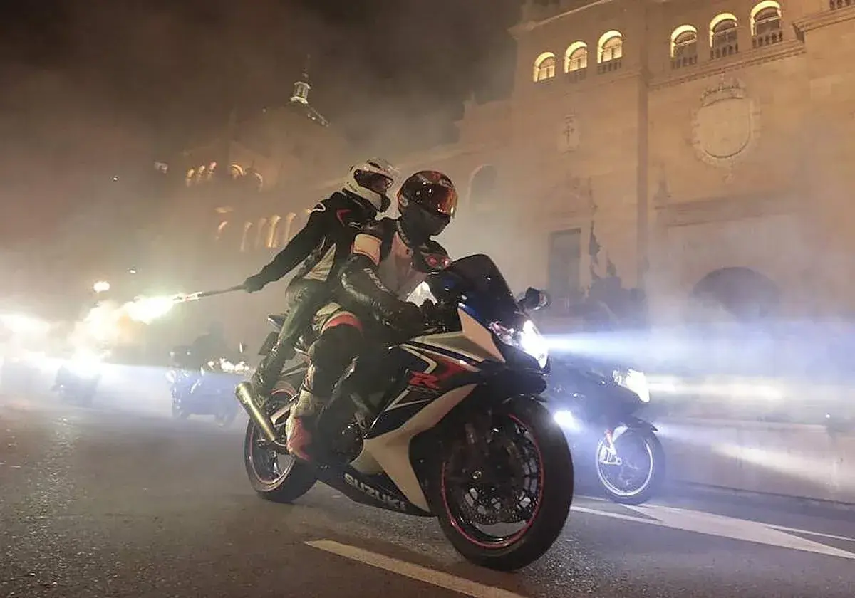 Dos motoristas en una Suzuki, uno con una bengala encendida, circulan por la noche. Parece que una motocicleta puede circular sin tubo de escape, creando un espectáculo de luces y humo.