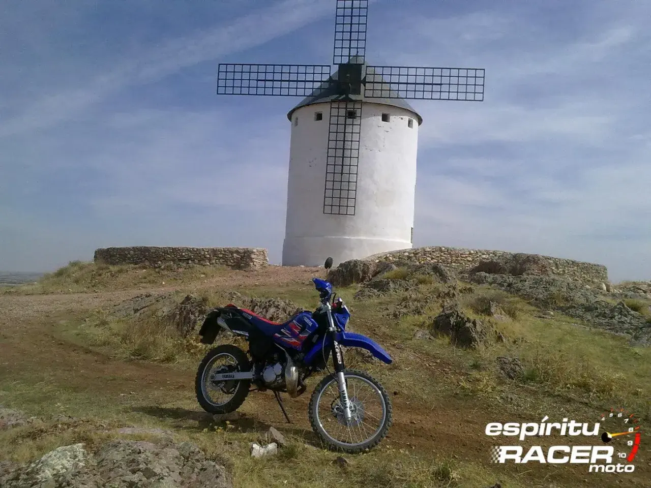 Yamaha DT 125 RE azul y roja aparcada junto a un molino de viento blanco en un paisaje rocoso.