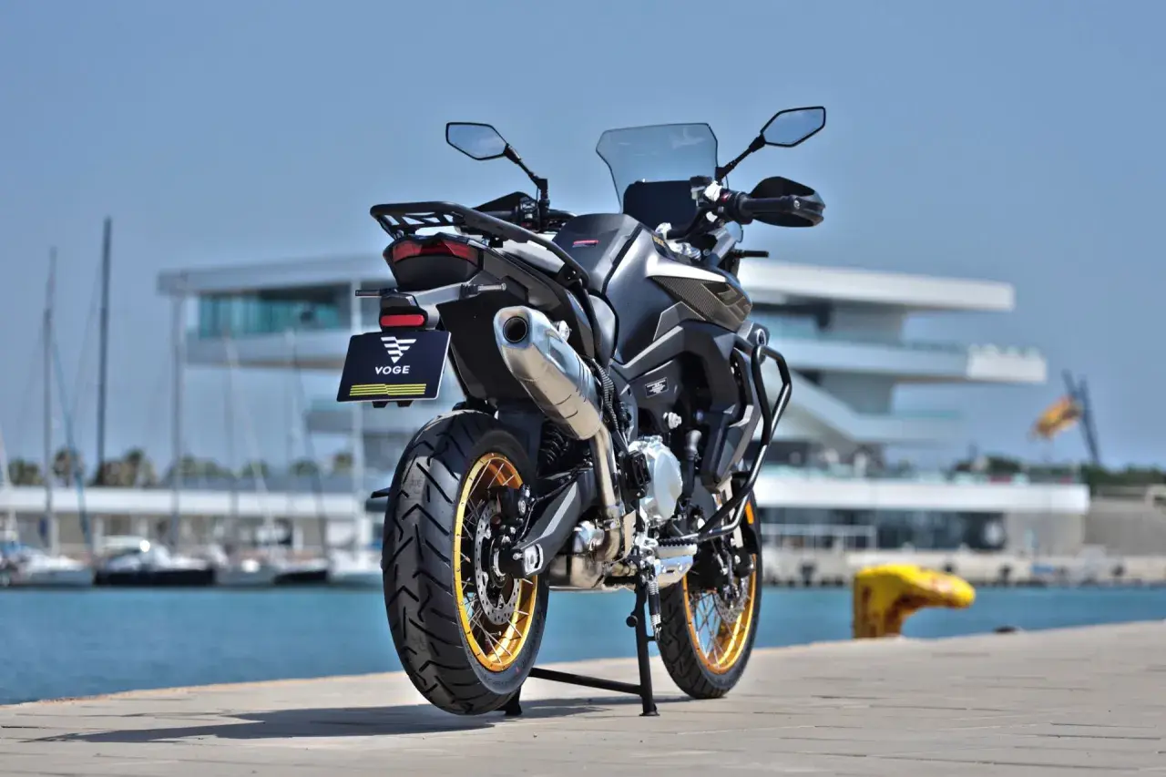 Voge 900 DSX ficha técnica: una moto de aventura negra con detalles dorados, aparcada junto a un puerto con veleros y un edificio moderno.