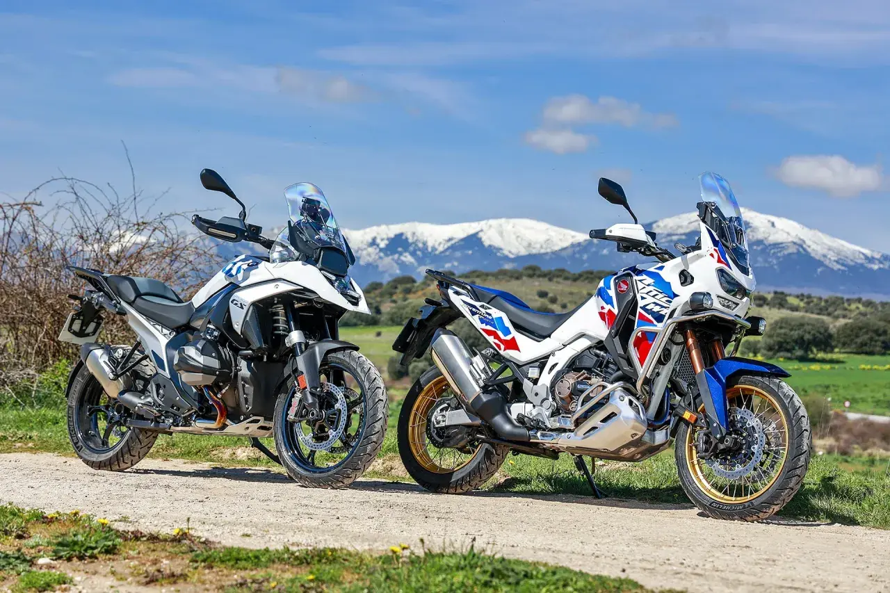 Dos motos de aventura, una Honda Africa Twin 2026 y otra blanca, aparcadas en un camino rural con montañas nevadas al fondo.