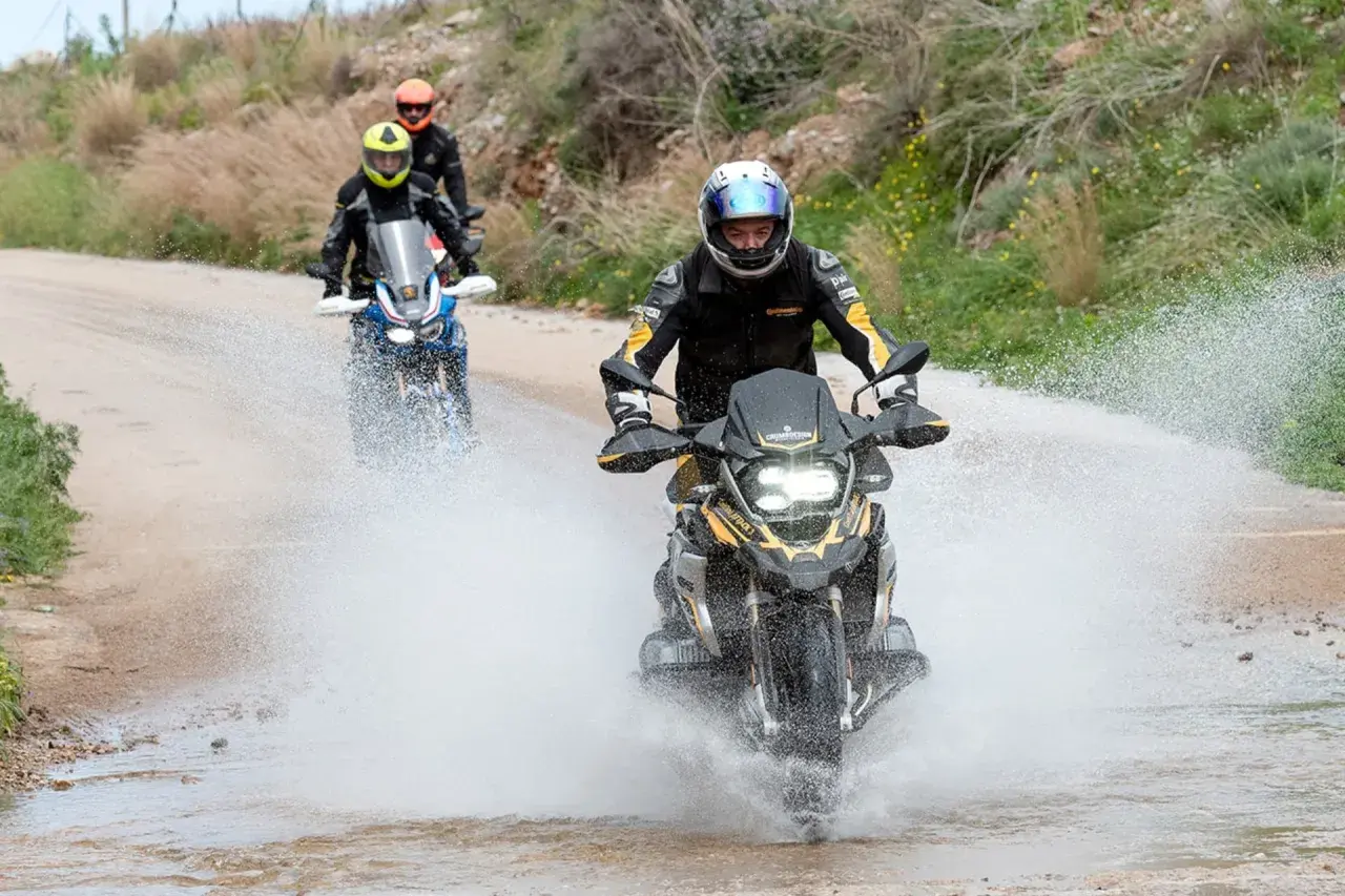 Motociclistas en un **conti trail attack 4 test** atraviesan un charco de agua en un camino de tierra, levantando una gran estela.