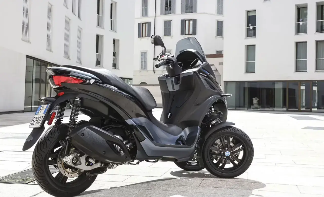Un Piaggio MP3 300 negro aparcado en una plaza urbana. Su diseño de tres ruedas ofrece estabilidad y un consumo eficiente.