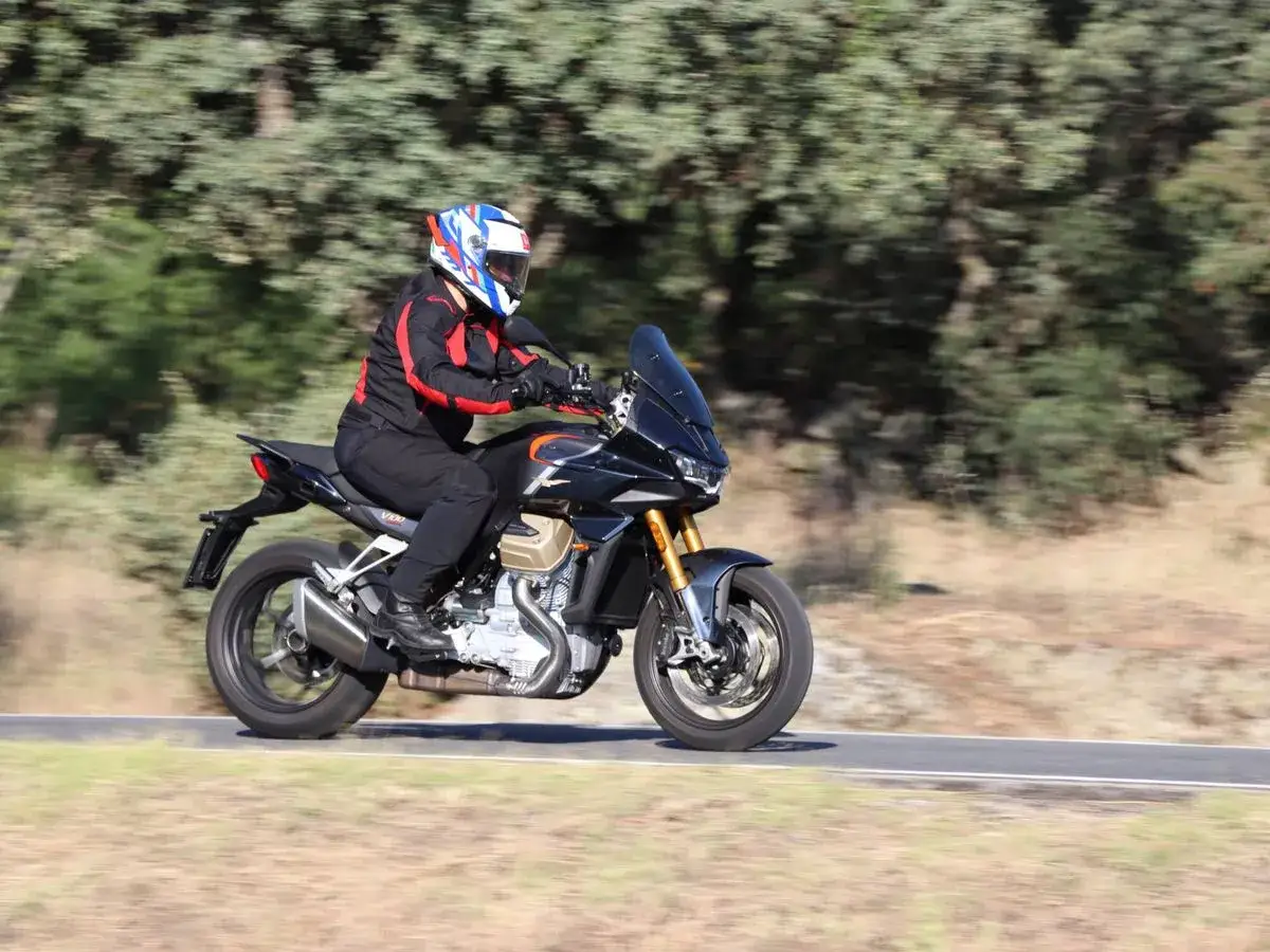 Motociclista en una Moto Guzzi Mandello negra, con casco y equipo de protección, circulando por una carretera rodeada de árboles.